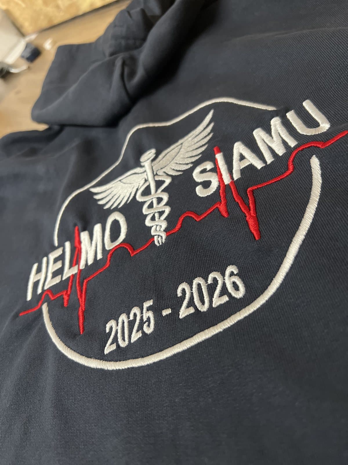 Broderie HELMo SIAMU 2025-2026 au dos d'un sweat marine